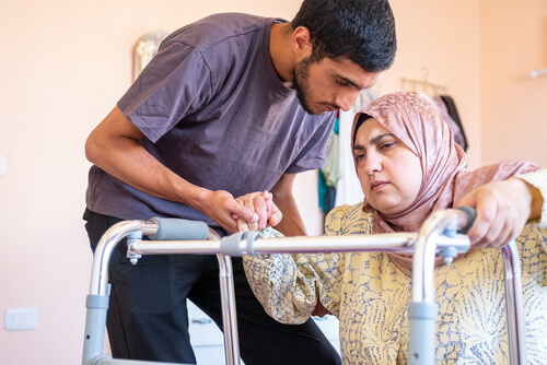 A young male caregiver assisting an older Muslim woman wearing a hijab as she uses a walking frame.