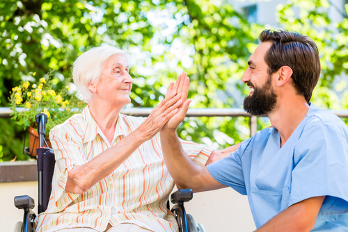 A male care worker in blue scrubs high-fiving an elderly woman sitting in a wheelchair outdoors in a garden setting.