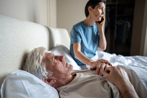A woman sitting on a bed speaking on the phone while holding the hand of an elderly man lying ill in bed.