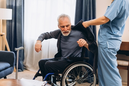 A caregiver in blue scrubs helping an elderly man in a wheelchair put on his jacket.