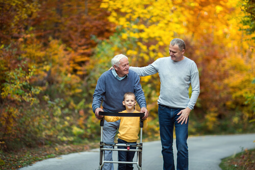 An elderly man using a walking frame walks along a tree-lined path in autumn with his adult son and young grandson.